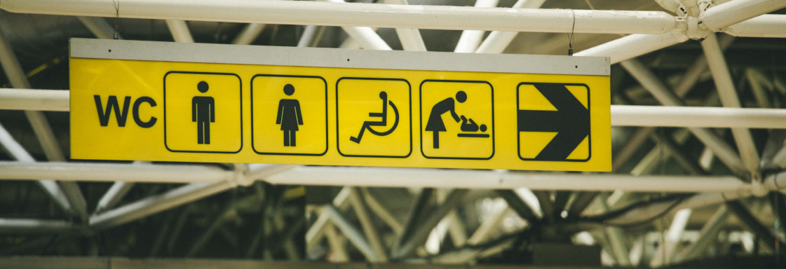 Imagen de señalización en un espacio público. De izquierda a derecha: WC, icono de hombre, icono de mujer, icono de persona usuaria de silla de ruedas, icono de cambiador de pañales, icono de una flecha señalando a la derecha. Foto por Paul Green para Unsplash
