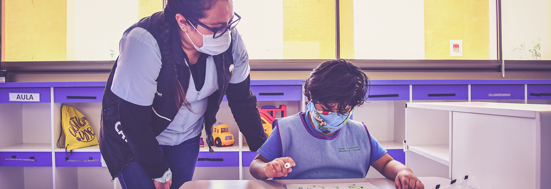 Fotografía de un niño frente a un pupitre de un salón de clases. Junto a él está una terapeuta. Se encuentran en el Centro Autismo Teletón.